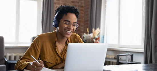 Woman sitting down at her desk writing her notes and talking to someone on her laptop